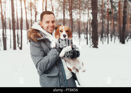 A young man dressed in a gray winter park in a snowy winter forest holds a dog named Beagle. Concept about winter leisure and love to animals Stock Photo