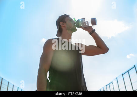 Waist up portrait of handsome young sportsman drinking water after workout outdoors standing against sky, lens flare Stock Photo