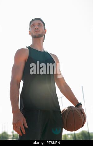 Low angle portrait of handsome young sportsman holding basketball ball  posing outdoors, lens flare Stock Photo