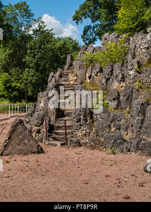 Fragment of the rock with its stairs knocked out in it and the grass and flowers with sharp peaks of stones in the park on a sunny day Stock Photo