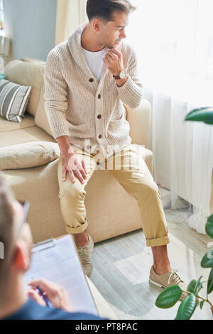 Serious thoughtful handsome young man in fashionable outfit focusing on his problem sitting on sofa handle and looking down while contemplating at the Stock Photo