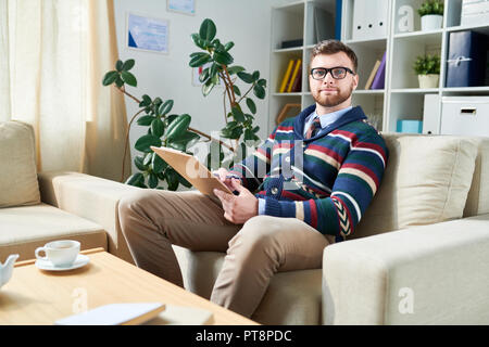 Content confident handsome young psychiatrist in glasses sitting in comfortable armchair and looking at camera while working in own office Stock Photo