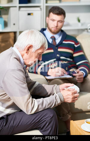 Serious pensive senior man sitting on sofa and looking down while telling about his fears with shrink, he holding napkin to wipe tears Stock Photo