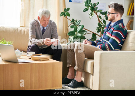 Serious senior man with napkin sitting on comfortable sofa and sharing his sufferings with shrink, concentrated psychiatrist analyzing words of patien Stock Photo