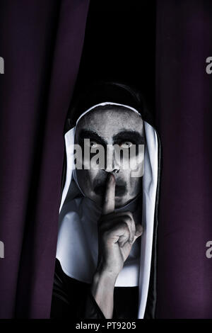 closeup of a frightening evil nun, wearing a typical black and white habit, peeping out from a purple curtain, asking for silence Stock Photo
