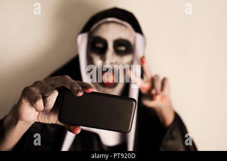 closeup of a frightening evil nun, wearing a typical black and white habit, taking a selfie with a smartphone while giving a V sign Stock Photo