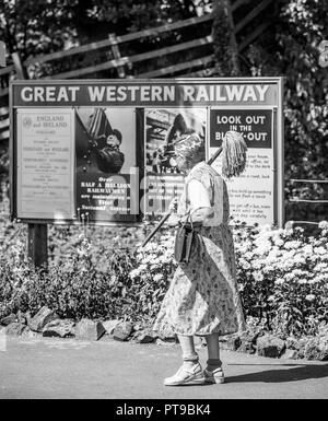 Monochrome rear of woman in overalls, headscarf, wrinkled stockings as vintage cleaning lady with mop at heritage railway station, UK 1940s WWII event. Stock Photo