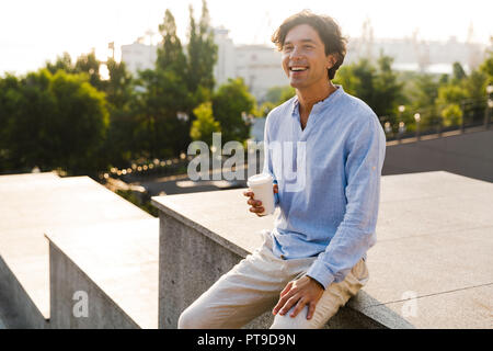Smiling man dressed in shirt drinking coffee while sitting outdoors at the city Stock Photo