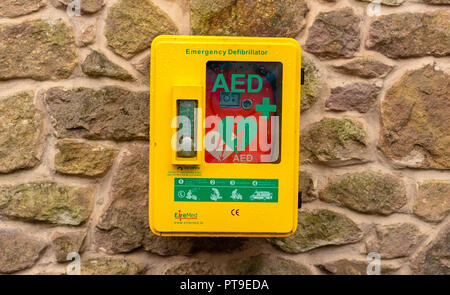Clean, new yellow and green Defibrillator hanging on large stone wall on a summers day in the UK, England Stock Photo