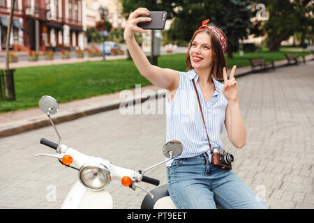 Happy young hipster woman making selfie on smartphone and showing peace gesture while sitting on scooter outdoors Stock Photo