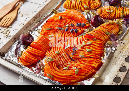 thinly sliced halves of pumpkins baked in an oven with onion, dried lavender, pumpkin seeds and spices on a rustic baking tray. wooden spatula on a wh Stock Photo