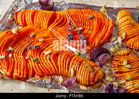 thinly sliced halves of pumpkins baked in an oven with onion, dried lavender, pumpkin seeds and spices on a rustic baking tray. wooden spatula on a wh Stock Photo