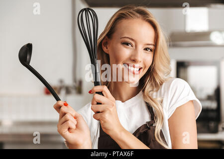 Smiling young woman chef cook in apron standing at the kitchen, holding ladle and whisk Stock Photo