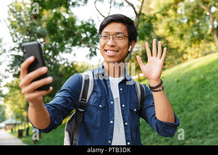 Happy asian male student in eyeglasses making selfie on smartphone and waving at camera while being in park Stock Photo