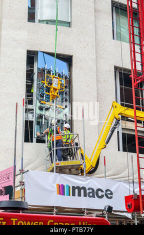 Building contractor Mace workers on a cherry picker hoist carefully fitting a large new glass window pane into a building in Mayfair, London, UK Stock Photo