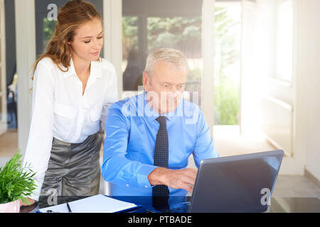 Shot of senior executive businessman sitting in front of laptop while young financial assitant businesswoman standing next to him and brainstorming to Stock Photo