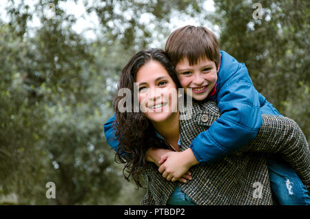Beautiful mother carrying blond hair son on her back outdoors. Concept of enjoying in family Stock Photo