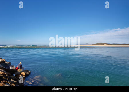 A man on his own wearing a hat sat on the boulders at the sea edge at Nambucca Heads, New South Wales, Australia Stock Photo