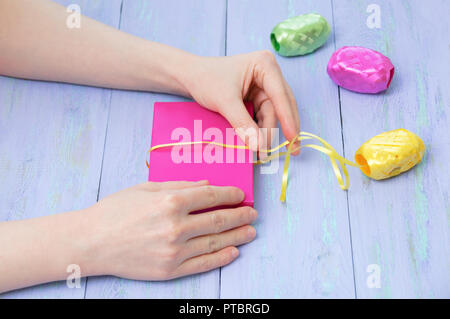Women'shands wrapped in a pink gift box with a yellow ribbon on a violet wooden background. Cose-up Stock Photo