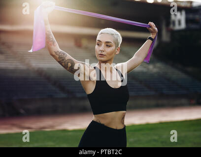 Woman runner standing in a track and field stadium doing stretching exercises holding a resistance band. Athletic woman in fitness clothes doing warm Stock Photo