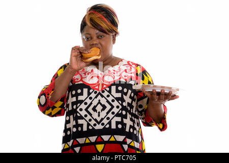 Studio shot of fat black African woman eating burger while holdi Stock Photo