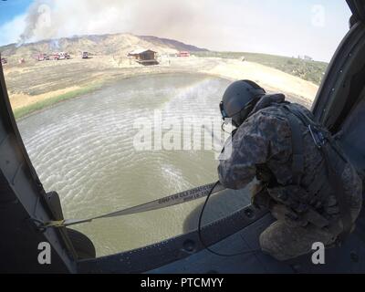 As a wildfire burns on a hill and California Department of Forestry and Fire Protection (CAL FIRE) vehicles and personnel stand by, Staff Sgt. Ge Xiong, crew chief aboard a UH-60 Black Hawk from the 1106th Theater Aviation Sustainment Maintenance Group (TASMG), California Army National Guard, watches a 2,600-gallon Bambi Bucket submerge into a pond during the Garza Fire in Kings County, California, July 13. The blaze consumed more than 26,000 acres and was 30 percent contained when this photo was taken. Five CalGuard Black Hawks responded to the Garza Fire, one of about two dozen torching Cali Stock Photo