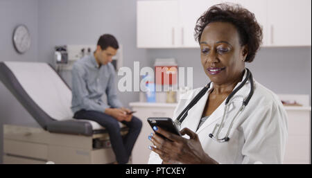 Middle aged African doctor using mobile phone to text while male patient waits Stock Photo