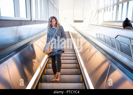 Low angle view, looking up of young woman standing on metro, subway, airport escalator going down with stairs, steps, bright light outside with people Stock Photo