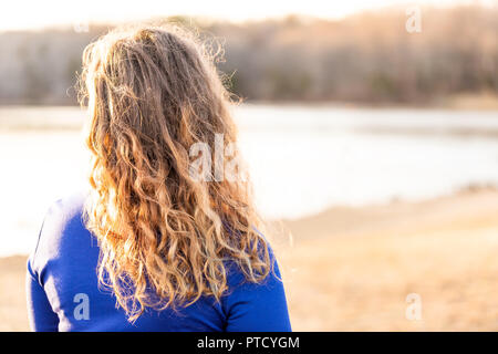Closeup back of young woman portrait standing by lake, river shore, water watching sunset, sunrise in blue dress behind Stock Photo