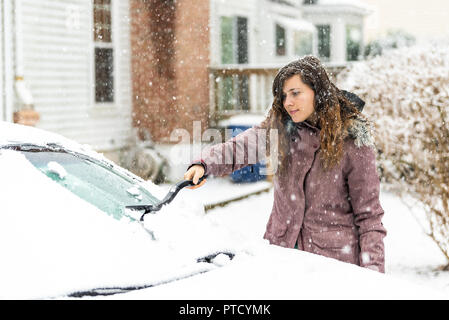 One young woman cleaning car windshield window from snow, ice with brush and scraper tool during snowfall while snowing snowflakes falling Stock Photo