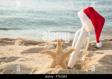 Santa hat on message in a bottle with starfish in beach sand and water background Stock Photo