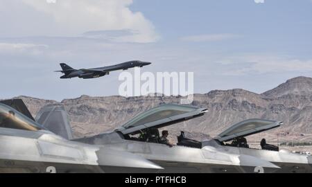 A group of F-22 Raptors from the 95th Fighter Squadron at Tyndall Air Force Base, Fla., sit on the flightline as a B-1B Lancer from the 34th Bomb Squadron at Ellsworth Air Force Base, S.D., takes off during Red Flag 17-3 at Nellis Air Force Base, Nev., July 18, 2017. Red Flag is designed to test the capability and interoperability between multiple air frames, and give the supporting personnel the chance to work together. Stock Photo