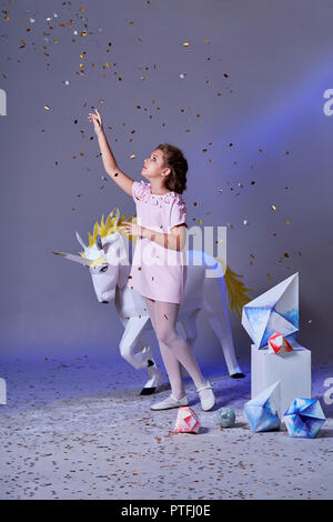 Holidays, christmas, new year, x-mas,concept. Little girl in elegance dress for party pointing finger on glitter confetti. Fashion lady teenage poses full-length in studio, gray purple background. Stock Photo