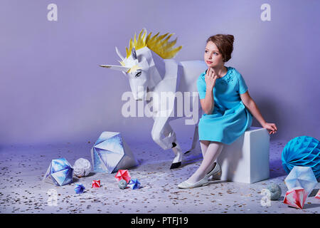 Romantic ,dreamy little girl lady origami studio, white big unicorn, geometric shapes. Beautiful,refined child in designer blue dress for holiday. Charming elegance teenager woman. Stock Photo