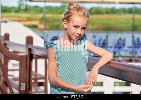 Portrait elegant young girl of 9-11 years outdoors, leaning her elbows on the railing. Cute child blonde smiling, sunny summer day resting on pier. Fashion kid concept. striped dress, marine style. Stock Photo