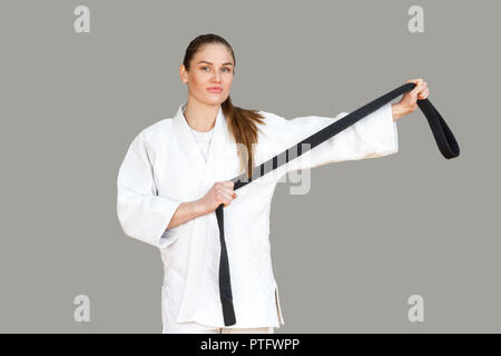 Confident attractive young athletic woman in white kimono standing and showing black belt, looking at camera. Japanese karate and sports concept. Indo Stock Photo