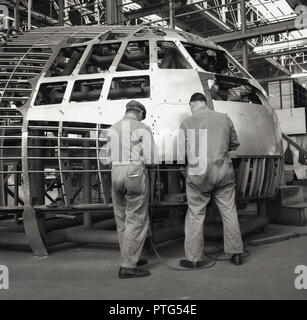 1950s, historical, aviation, two male mechanics or engineers in overalls working on the exterior of an aircraft cockpit inside an aerospace factory or hanger, England, UK. Stock Photo