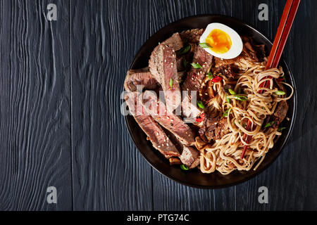 black bowl of Soba noodles with sliced roast beef, shiitake mushrooms, half of hard boiled egg and fried vegetables with chopsticks, asian cuisine, vi Stock Photo