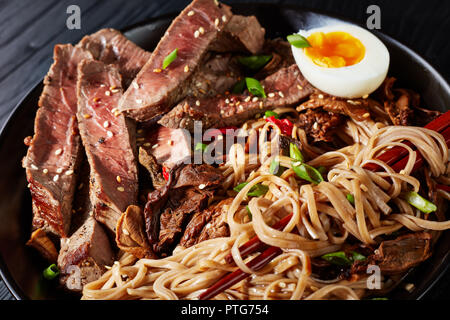 close-up of black bowl of Soba noodles with sliced roast beef, shiitake mushrooms, half of hard boiled egg and fried vegetables with chopsticks, asian Stock Photo