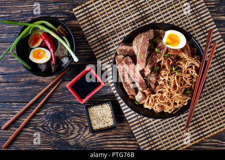 a bowl of soba with sliced roast beef, shiitake mushrooms, half of boiled egg and vegetables with chopsticks on a bamboo table mat. ingredients on a r Stock Photo