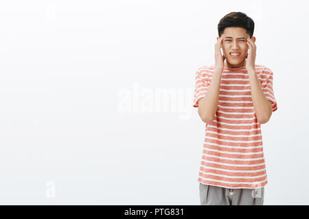 Young uneasy asian guy in striped t-shirt feeling pressured and tired holding hands on temples, suffering headache or migraine, clenching teeth from pain and intense thinking over grey wall Stock Photo