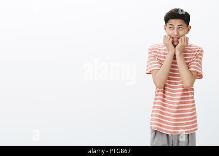 Portrait of nervous and scared young asian boy in striped t-shirt biting fingers looking at upper left corner insecure and concerned, feeling frightened and worried over white wall Stock Photo