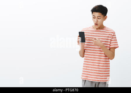 Portrait of impressed and surprised excited young attractive chinese guy in striped t-shirt open mouth from interest and thrill holding smartphone pointing and looking at cellphone screen fascinated Stock Photo