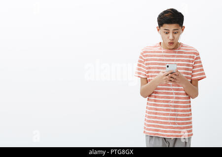 Impressed happy excited attractive young asian male with cool hairstyle in striped t-shirt holding smartphone looking pleased and amazed at cellphone screen saying wow, posing over white wall Stock Photo