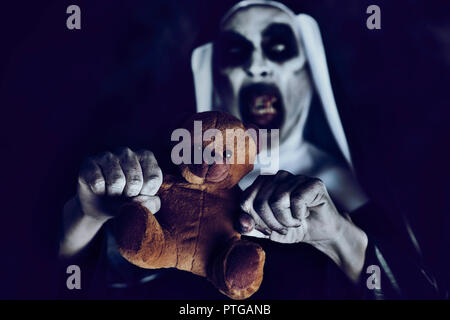 closeup of a frightening evil nun, with bloody teeth and scary eyes, wearing a typical black and white habit, trying to dismember a teddy bear against Stock Photo