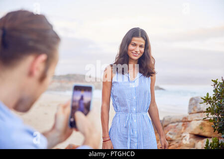 Young man with smart phone photographing girlfriend at beach Stock Photo