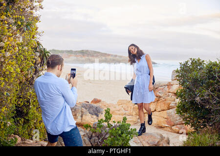 Young man with smart phone photographing girlfriend with ocean in background Stock Photo