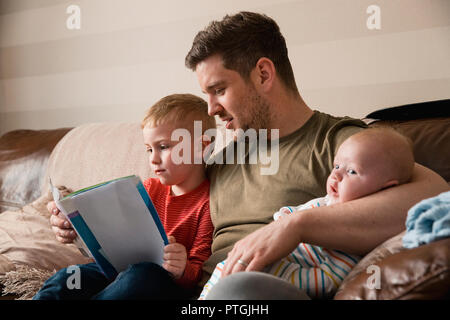 Mid-adult father is holding his newborn baby boy in his arm and then reading though the book with his son under the other arm. They all all sitting on Stock Photo