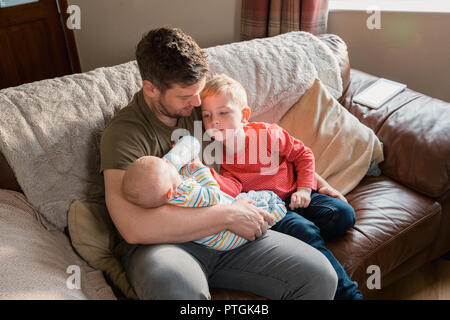 Father sitting on the sofa in the living room with his newborn baby boy in his arms giving him a bottle of milk and his other son sitting next to him  Stock Photo