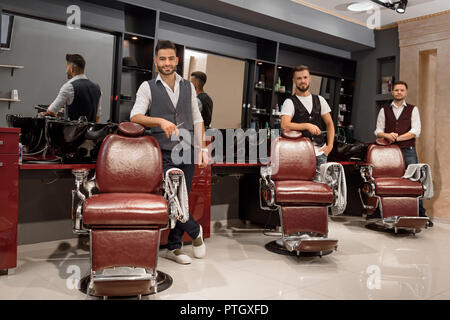 Handsome, confident masters looking at camera and holding scissors in hands. Three professional barbers standing near hairdresser chairs and posing. Interior of barbershop. Stock Photo